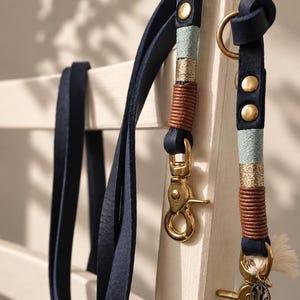 Pode incluir: Uma trela de couro azul-escuro com mosquetões e argolas de metal dourado. A trela é decorada com fios em azul-claro, dourado metalizado e um cordão de couro castanho, incluindo um pequeno pendente de metal em forma de âncora.
