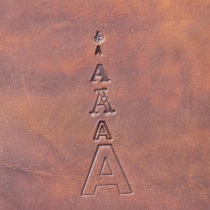 May include: A brown leather surface featuring a vertical column of five stamped letter A characters. The letters vary in size and font style, arranged from smallest at the top to largest at the bottom, with a small cross symbol above the top letter.
