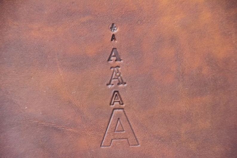 May include: A brown leather surface featuring a vertical column of five stamped letter A characters. The letters vary in size and font style, arranged from smallest at the top to largest at the bottom, with a small cross symbol above the top letter.