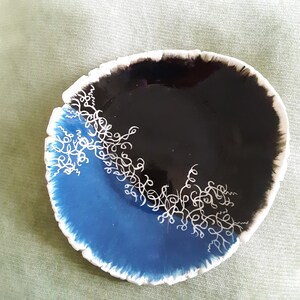 Puede incluir: Un plato pequeño y redondo de cerámica con un esmalte azul y negro. El plato tiene un diseño blanco de líneas en espiral en el borde.