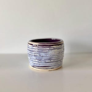 May include: A small, cylindrical ceramic pot featuring a deep purple interior and a textured, ribbed exterior. The outer surface displays a layered finish with white glaze over a dark purple base, creating a distressed, horizontal striped appearance.