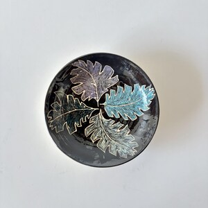 May include: A round ceramic bowl with a glossy black glaze featuring three distinct leaf impressions in the center. The leaves are colored in shades of teal, light blue, and muted purple, each outlined with a fine, light-colored etched detail.