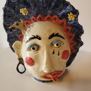May include: A ceramic wall mask featuring a pale face with red cheeks, blue eyeshadow, and a tongue sticking out. The head is framed by a large, ruffled blue collar with a red serrated trim, small yellow floral accents, and a small bird figure.