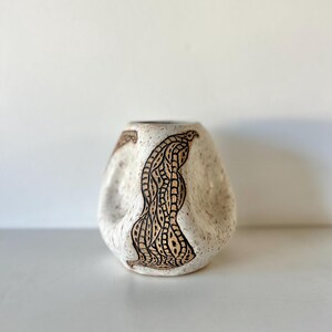 May include: A ceramic vase with a speckled off-white finish and a distinct, irregular, pinched shape. The surface features dark, intricate, hand-drawn linear patterns that follow the curves of the vessel, creating a textured, organic appearance.