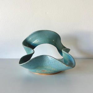 May include: A ceramic bowl with a wavy, ribbon-like rim and a speckled light blue glaze. The base is circular and flat, showing a natural brown clay color. The interior and exterior surfaces feature a smooth, glossy finish with dark speckles.