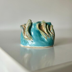 May include: A small, round ceramic vessel with a glossy turquoise glaze. The exterior features raised, organic, wave-like protrusions with a textured, sandy-brown finish along the edges, contrasting with the smooth, shiny blue surface.