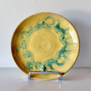 May include: A round ceramic plate featuring a vibrant yellow glaze with a decorative ring of teal, fern-like patterns radiating from the center. The surface has a glossy finish with subtle texture and small speckles throughout the yellow base.
