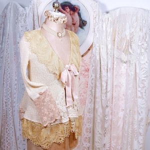 May include: A cream-colored crocheted cardigan with a lace trim and a pink satin ribbon tied at the neckline. The cardigan is layered over a white lace top and is displayed on a mannequin.