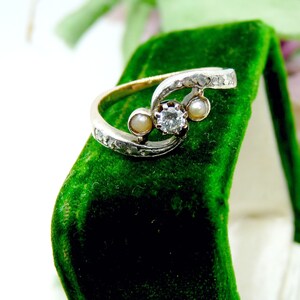 May include: A silver ring with a diamond and two pearls set in a swirling design. The ring is displayed in a green velvet ring box.