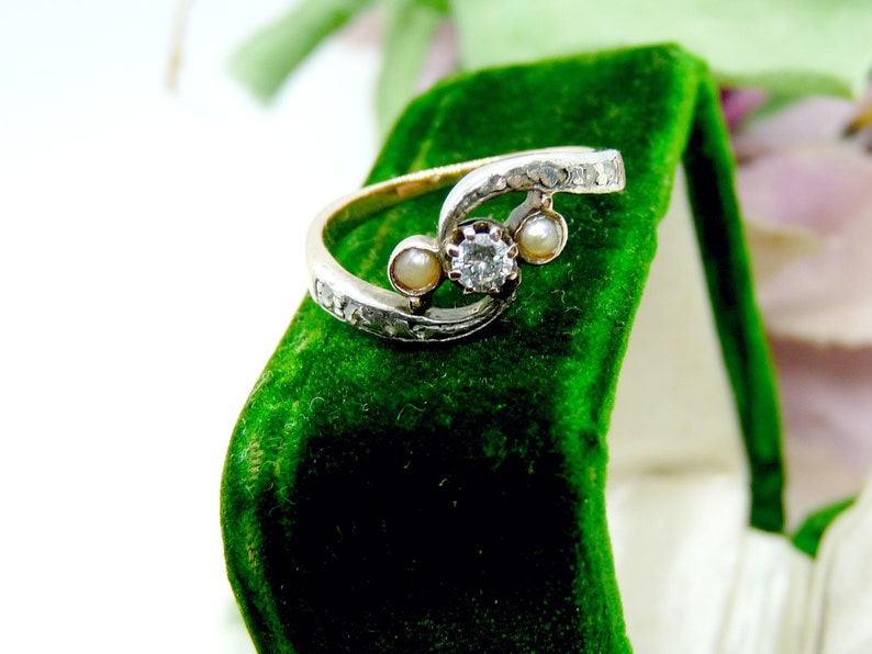 May include: A silver ring with a diamond and two pearls set in a swirling design. The ring is displayed in a green velvet ring box.