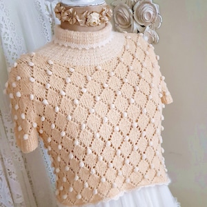 Peut inclure: Un haut en crochet de couleur crème avec un col montant et des manches courtes. Le haut présente un motif en forme de diamant avec des boules blanches partout.