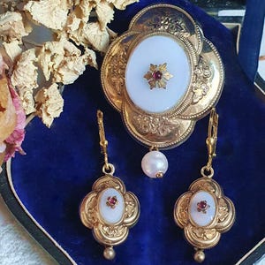 Op de afbeelding: Een vintage sieradenset met een goudkleurige broche en bijpassende oorbellen. De broche heeft een ovale witte kern met een rode edelsteen en een pareldruppel. De oorbellen hebben ook witte ovale kernen met rode edelstenen.