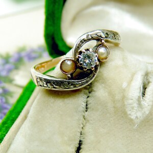 May include: A silver ring with a diamond and two pearls set in a swirling design. The ring is in a white velvet box.
