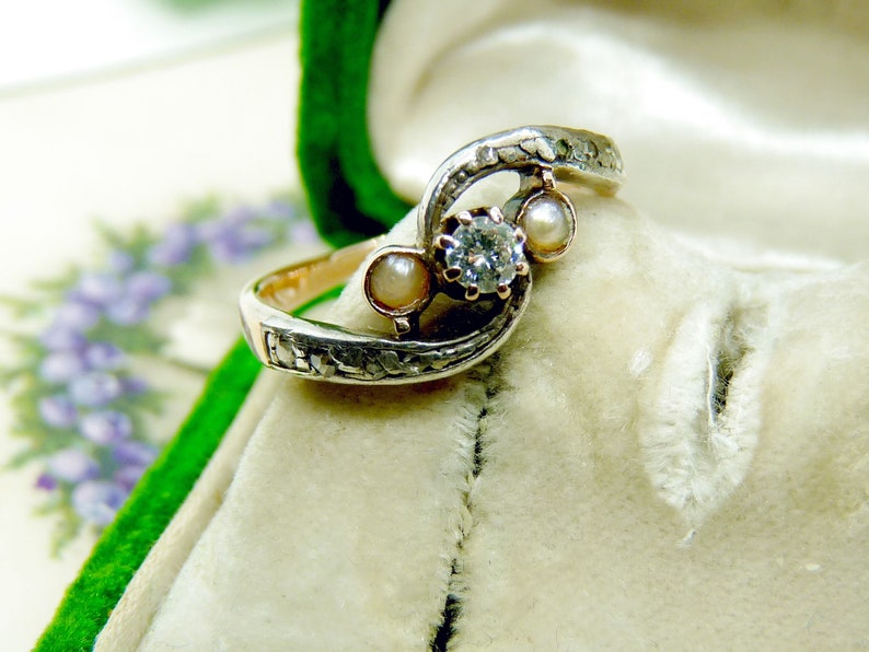 May include: A silver ring with a diamond and two pearls set in a swirling design. The ring is in a white velvet box.