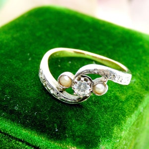 May include: A silver ring with a diamond and two pearls set in a swirling design. The ring is on a green velvet background.