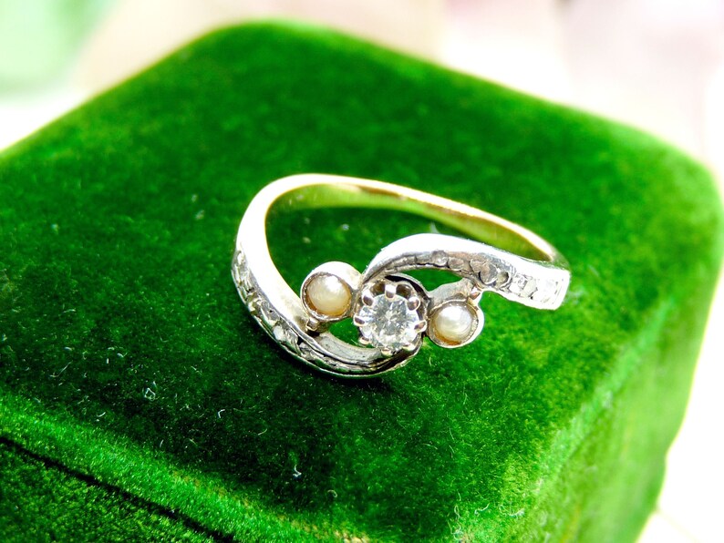 May include: A silver ring with a diamond and two pearls set in a swirling design. The ring is on a green velvet background.