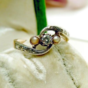 May include: A silver and gold ring with a diamond and two pearls. The ring is set in a delicate, swirling design.