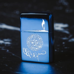 May include: A blue metal lighter with an engraved design of a hand holding a circle with various symbols. The symbols include a tree, a drop of water, a star, and a crescent moon.