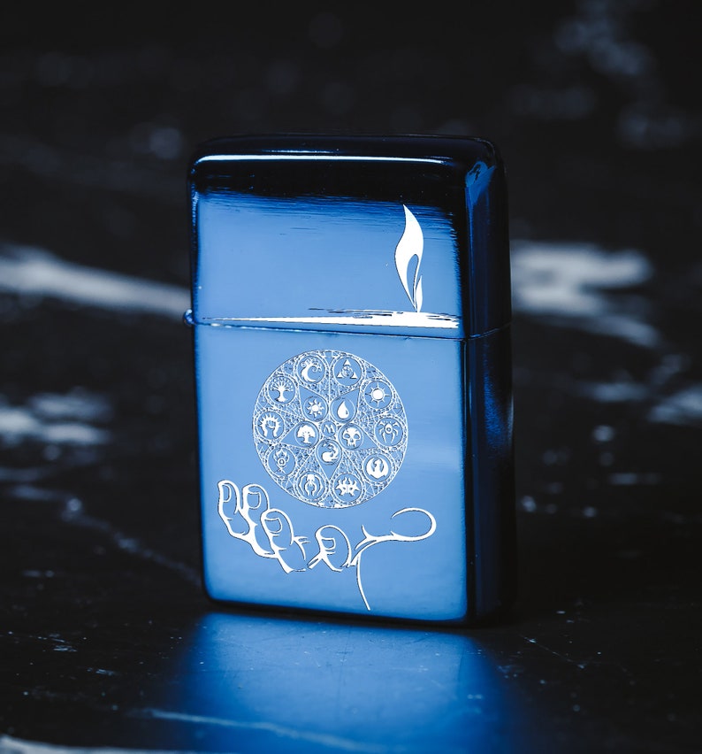 May include: A blue metal lighter with an engraved design of a hand holding a circle with various symbols. The symbols include a tree, a drop of water, a star, and a crescent moon.