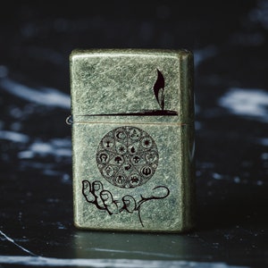 May include: A vintage-style lighter with a green, textured finish. The lighter features a hand holding a circle with various symbols, including a star, a crescent moon, and a sun.