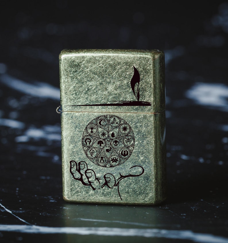 May include: A vintage-style lighter with a green, textured finish. The lighter features a hand holding a circle with various symbols, including a star, a crescent moon, and a sun.