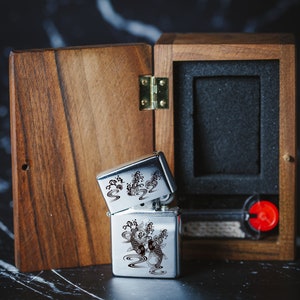 May include: A silver lighter featuring a black and white design of koi fish and waves. The lighter is presented in a wooden box with a black interior.