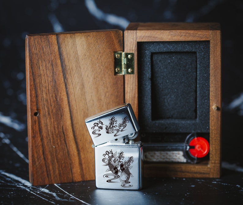 May include: A silver lighter featuring a black and white design of koi fish and waves. The lighter is presented in a wooden box with a black interior.