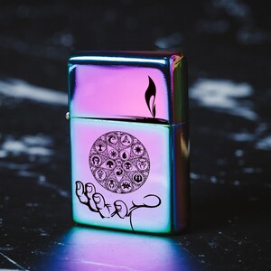 May include: A iridescent metal lighter with a black and white design of a circle with symbols inside. The lighter has a black flame design on the top.