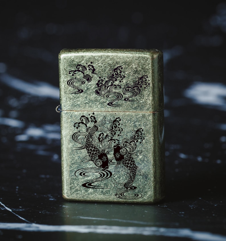 May include: A vintage-style lighter with a green metallic finish and an engraved design of two black koi fish swimming in a wave pattern.