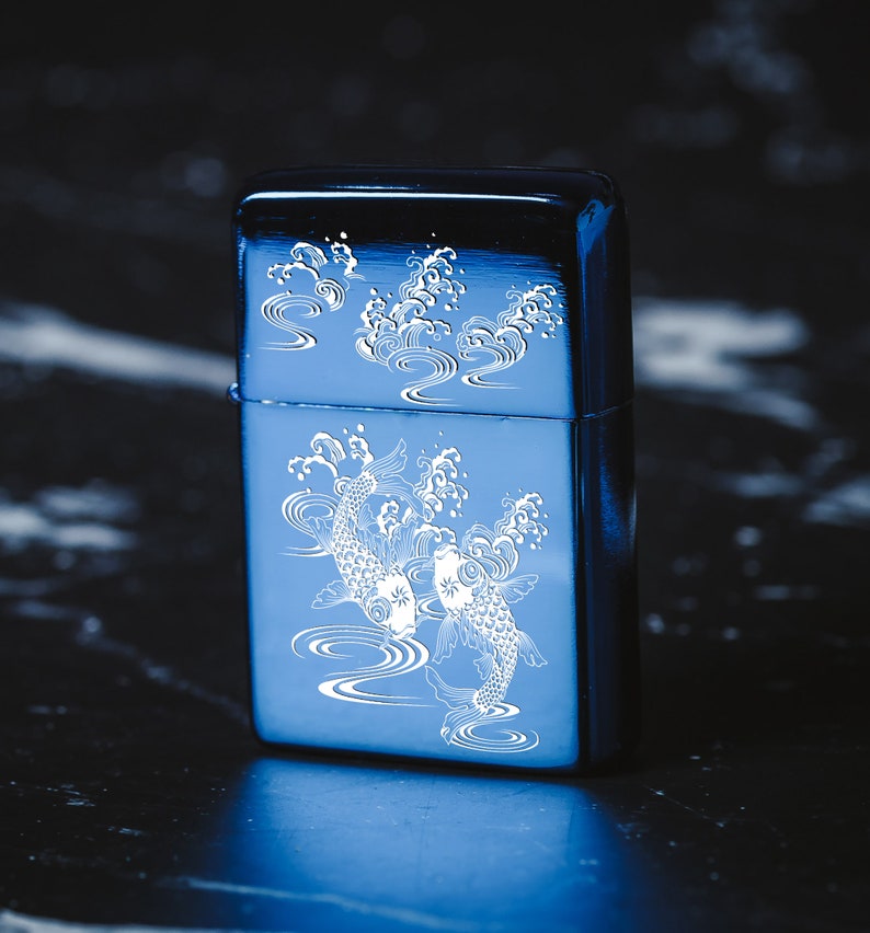 May include: A blue metal lighter featuring a white design of two koi fish swimming in a wave pattern.