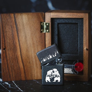 Puede incluir: Un encendedor negro con un dise&ntilde;o blanco de dos osos y &aacute;rboles dentro de una caja de madera. La caja es de madera oscura con una tapa con bisagras.