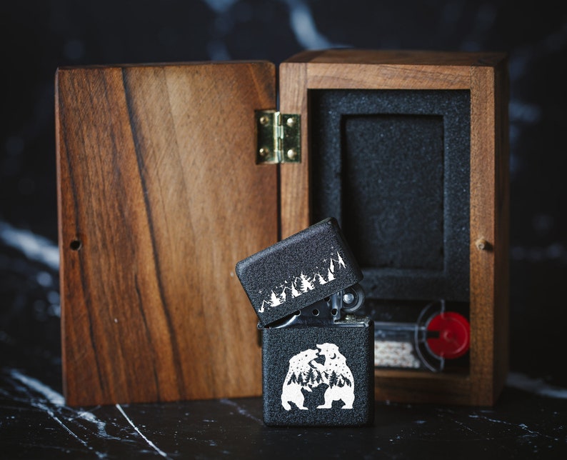 Puede incluir: Un encendedor negro con un dise&ntilde;o blanco de dos osos y &aacute;rboles dentro de una caja de madera. La caja es de madera oscura con una tapa con bisagras.