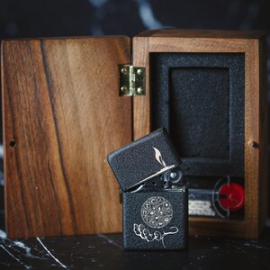 May include: A black lighter with a silver hinge and a design of a circle with symbols inside. The lighter is open and is sitting in a wooden box with a black interior.