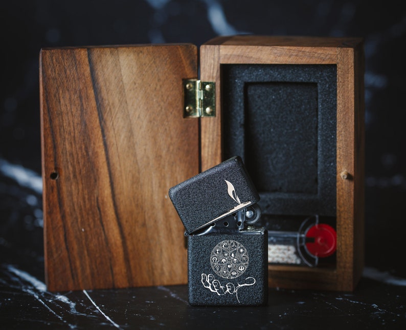May include: A black lighter with a silver hinge and a design of a circle with symbols inside. The lighter is open and is sitting in a wooden box with a black interior.