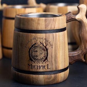 May include: A wooden beer stein with a black band around the top and bottom. The stein has a stainless steel interior and a carved wooden handle. The front of the stein features a wood-burned image of a hobbit hole with the text "In a hole in the ground there lived a hobbit" and "Home is a hobbit hole". The name "MICHAEL" is also engraved on the stein.