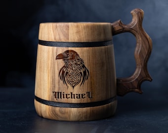 May include: Wooden beer mug with a carved raven design and the name "Michael" in a gothic font. The mug has a dark wood handle and is accented with black bands. The raven is intricately detailed with Celtic knotwork.