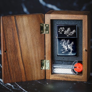May include: A wooden box with a hinged lid, open to reveal a black lighter with a white design of koi fish and water. The lighter is in a black and white box with a white border. The box is on a black and white marble surface.