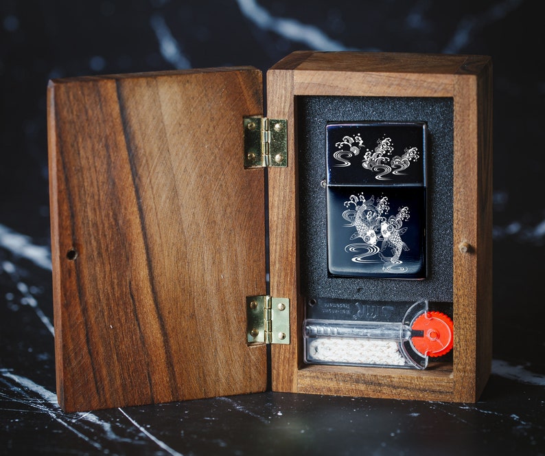 May include: A wooden box with a hinged lid, open to reveal a black lighter with a white design of koi fish and water. The lighter is in a black and white box with a white border. The box is on a black and white marble surface.