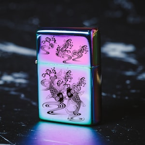 May include: A silver lighter featuring a black and white design of two koi fish swimming in a wave pattern. The lighter has a rainbow iridescent finish.