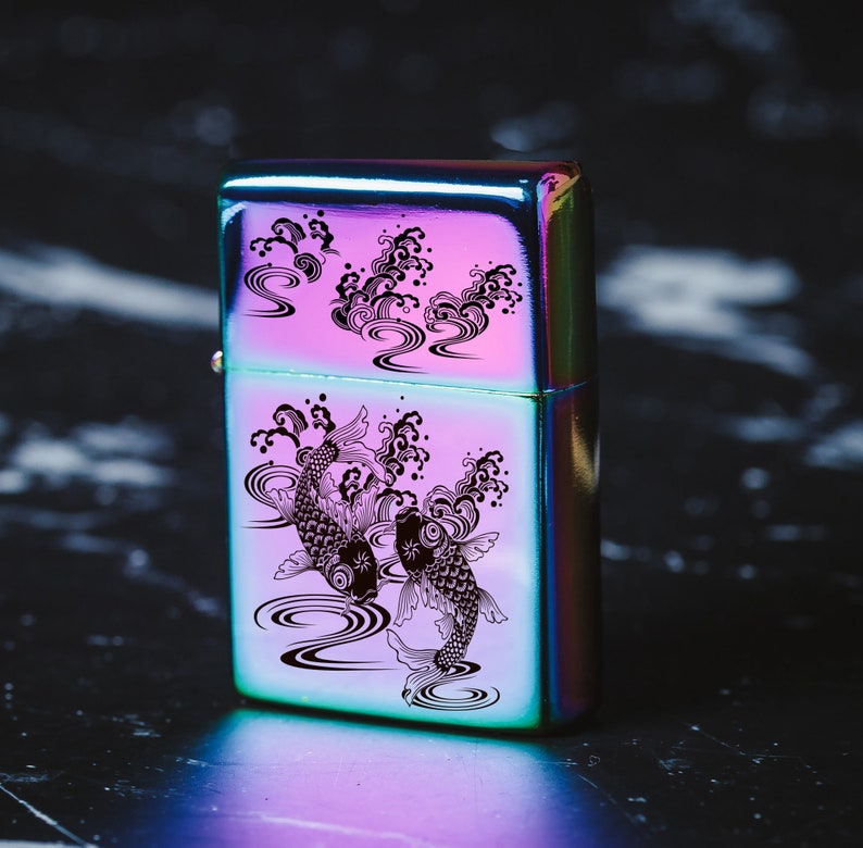 May include: A silver lighter featuring a black and white design of two koi fish swimming in a wave pattern. The lighter has a rainbow iridescent finish.