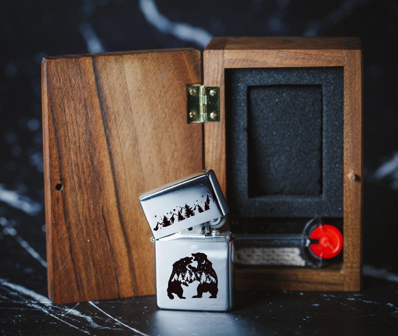 Puede incluir: Un encendedor plateado con un dise&ntilde;o en blanco y negro de dos osos luchando en un bosque. El encendedor est&aacute; en una caja de madera con un interior negro.