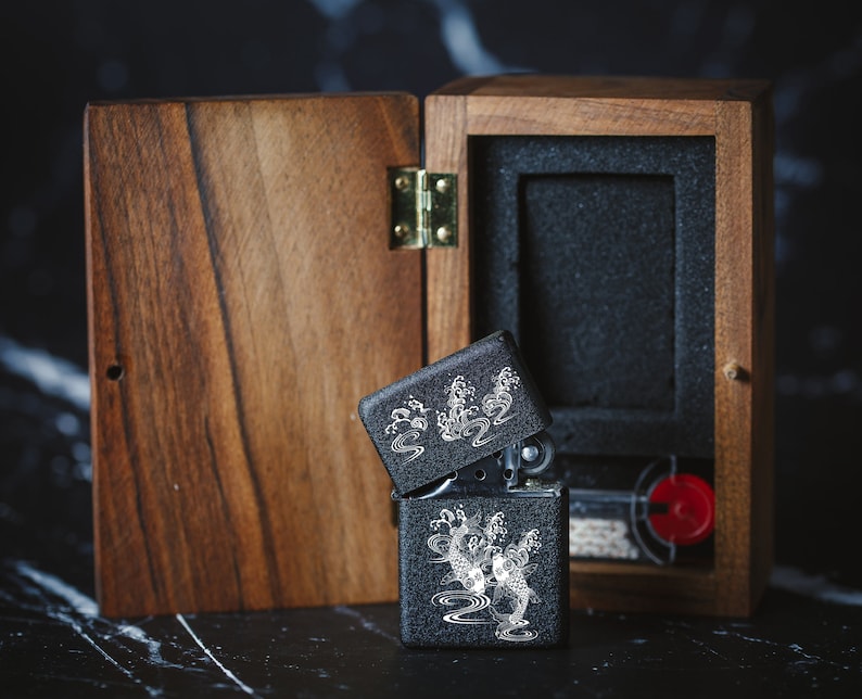 May include: A black lighter with a silver design of koi fish and waves in a wooden box. The box is open and the lighter is partially out of the box.