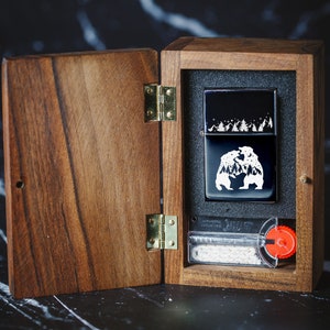 Puede incluir: Una caja de madera con una tapa con bisagras, abierta para revelar un encendedor negro con un dise&ntilde;o blanco de dos osos y &aacute;rboles. El encendedor est&aacute; en un forro de fieltro negro. La caja tiene una bisagra de lat&oacute;n y una perilla roja.