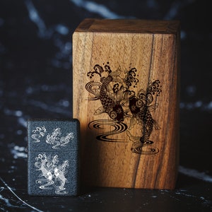 May include: A black wooden box with a decorative design of two koi fish swimming in a stream. The box is next to a black lighter with a similar design.