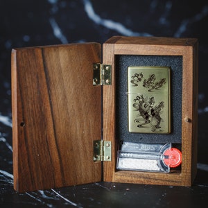 May include: A wooden box with a hinged lid, open to reveal a gold lighter with an engraved design of koi fish swimming in water. The lighter is in a black velvet-lined compartment. The box has a brass hinge and a small compartment with a white wick and a red knob.
