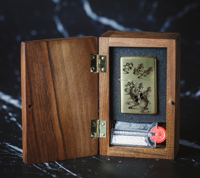 May include: A wooden box with a hinged lid, open to reveal a gold lighter with an engraved design of koi fish swimming in water. The lighter is in a black velvet-lined compartment. The box has a brass hinge and a small compartment with a white wick and a red knob.