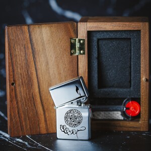 May include: A silver lighter with an engraved design of a circle with symbols inside and a hand underneath. The lighter is in a wooden box with a hinged lid.