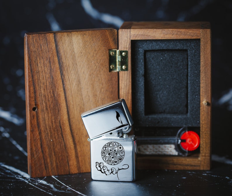 May include: A silver lighter with an engraved design of a circle with symbols inside and a hand underneath. The lighter is in a wooden box with a hinged lid.