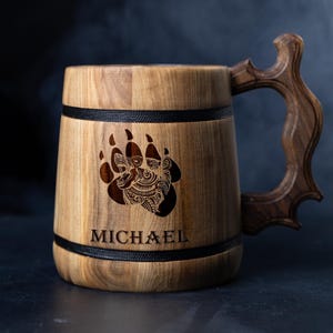 Könnte beinhalten: Ein Bierkrug aus Holz mit einem schwarzen Lederband und einem Bärenpfotenabdruck. Der Krug hat einen geschnitzten Holzgriff und der Name "MICHAEL" ist auf der Vorderseite eingraviert.