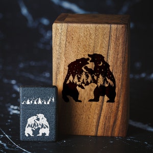 Puede incluir: Una caja de madera negra con un dise&ntilde;o grabado con l&aacute;ser de dos osos que se enfrentan. Los osos est&aacute;n delineados en blanco y tienen una escena de bosque dentro de ellos. La caja est&aacute; al lado de un encendedor negro con un dise&ntilde;o similar.